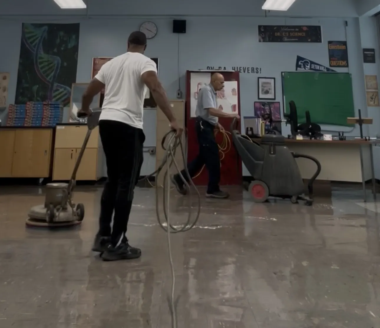 Two TLC crew members working a classroom floor with a rotary floor machine and a wet vacuum.